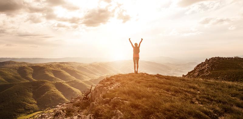 image of a young woman raising her hands in triumph on a tall cliff facing the sunset