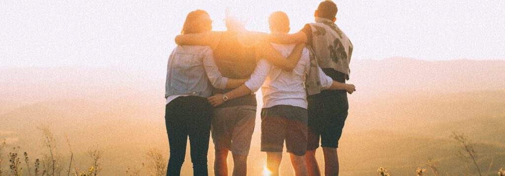 four person hands wrap around shoulders while looking at sunset