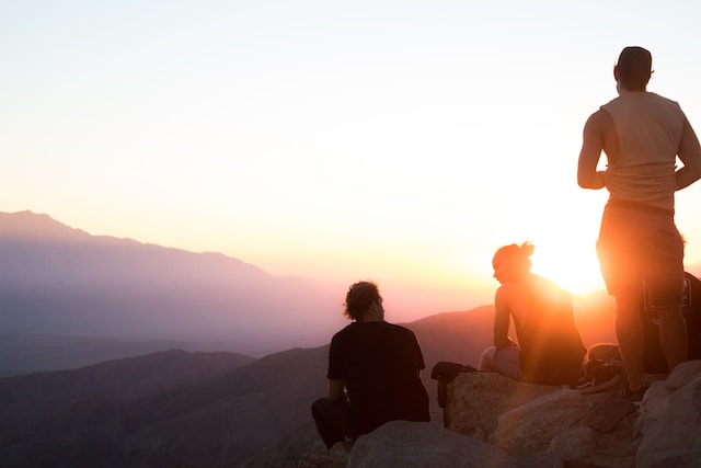 three people sitting on a hill at sunset