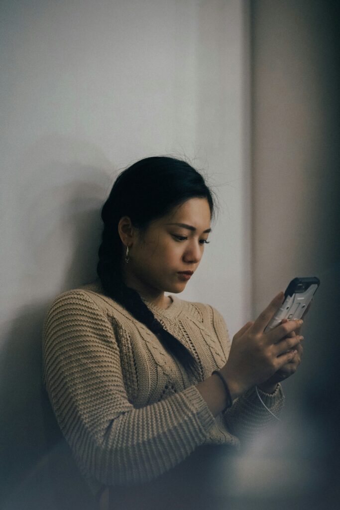 woman sitting against a wall looking at her phone dejectedly 