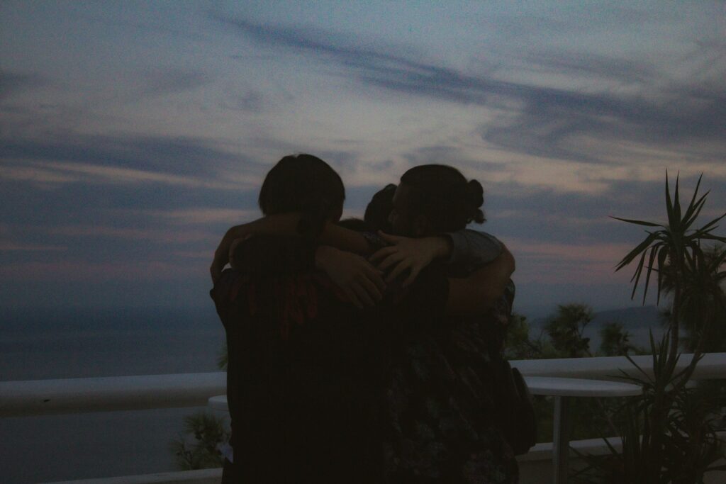 three people hugging from behind, it appears to be on a deck near the ocean in very dim light