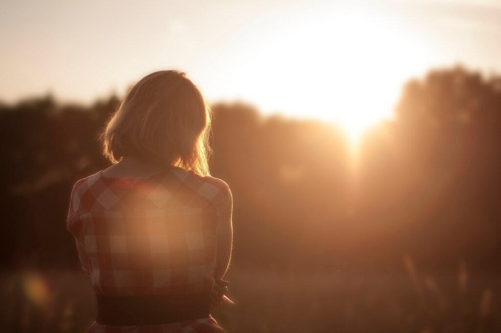 a woman looking at a sunset, from behind, the sun is about to dip below trees