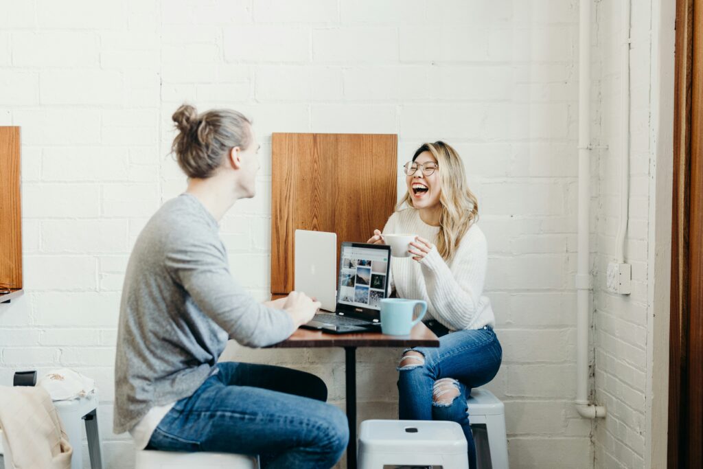 Two people sitting across from eachother at a small coffee shop table. A woman is facing and laughing and a man is seen from behind.