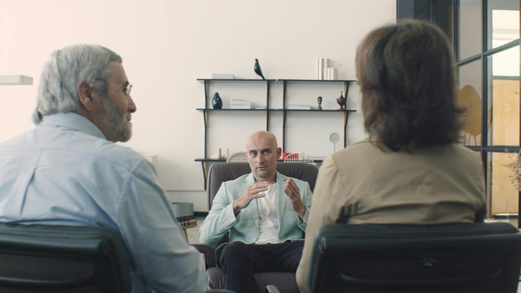 Two adults sitting in front of a desk talking with a bald man