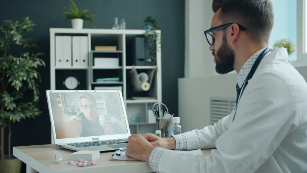 A telehealth doctor addresses a patient on a screen.