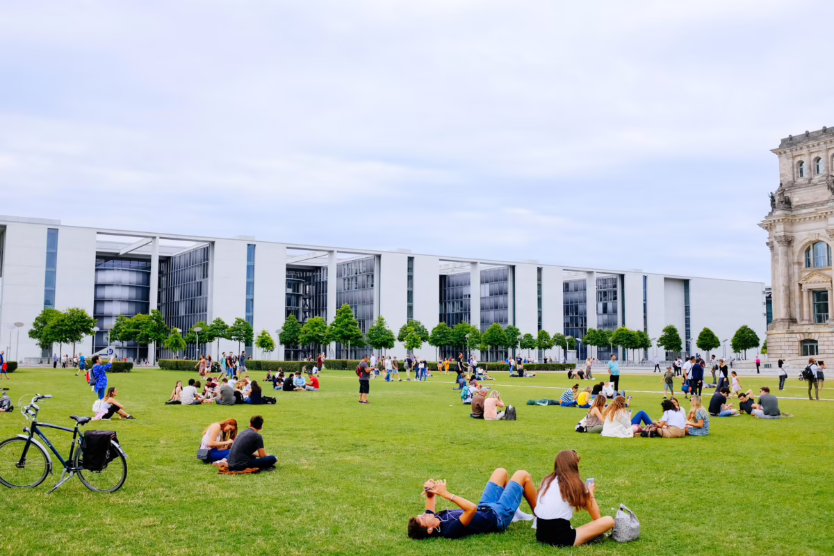 Many small groups of young adults sit and walk on a large, grassy field with trees and buildings surrounding it. A bike and other modes of transportation are present