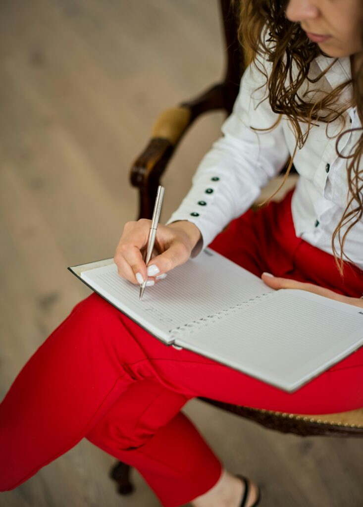 A woman wearing a white jacket and red pants sits and writes in a notebook
