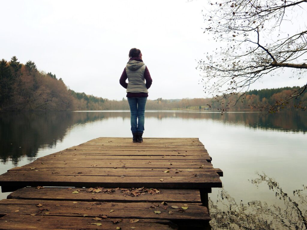 A woman in jeans and a vest with long sleeve shirt stands on a dock looking out onto a body of water